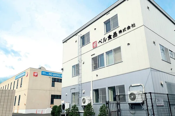 青空の下に立つ白い社屋。壁に「ベル食品株式会社」の社名とロゴが掲げられ、奥にISO22000認証の表示がある建物が並ぶ