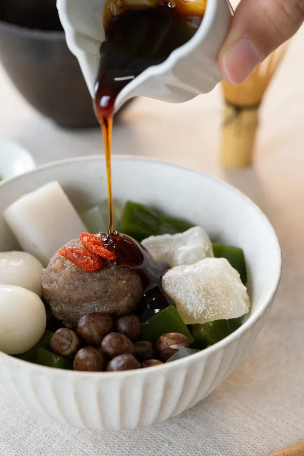 器のあんみつに、白い片口から黒蜜を注いでいるところ。奥に抹茶の茶碗と茶筅