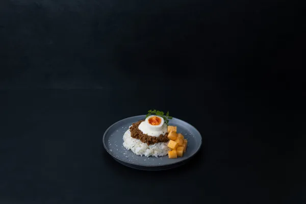 黒い皿に盛られたキーマカレー。半熟卵と角切りのメロンが添えられている