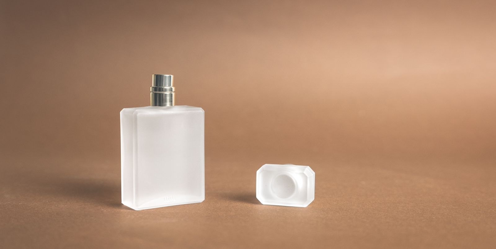 茶色の背景に置かれた淡いガラスの香水瓶とキャップ。本文で扱う製品とは異なるイメージ写真