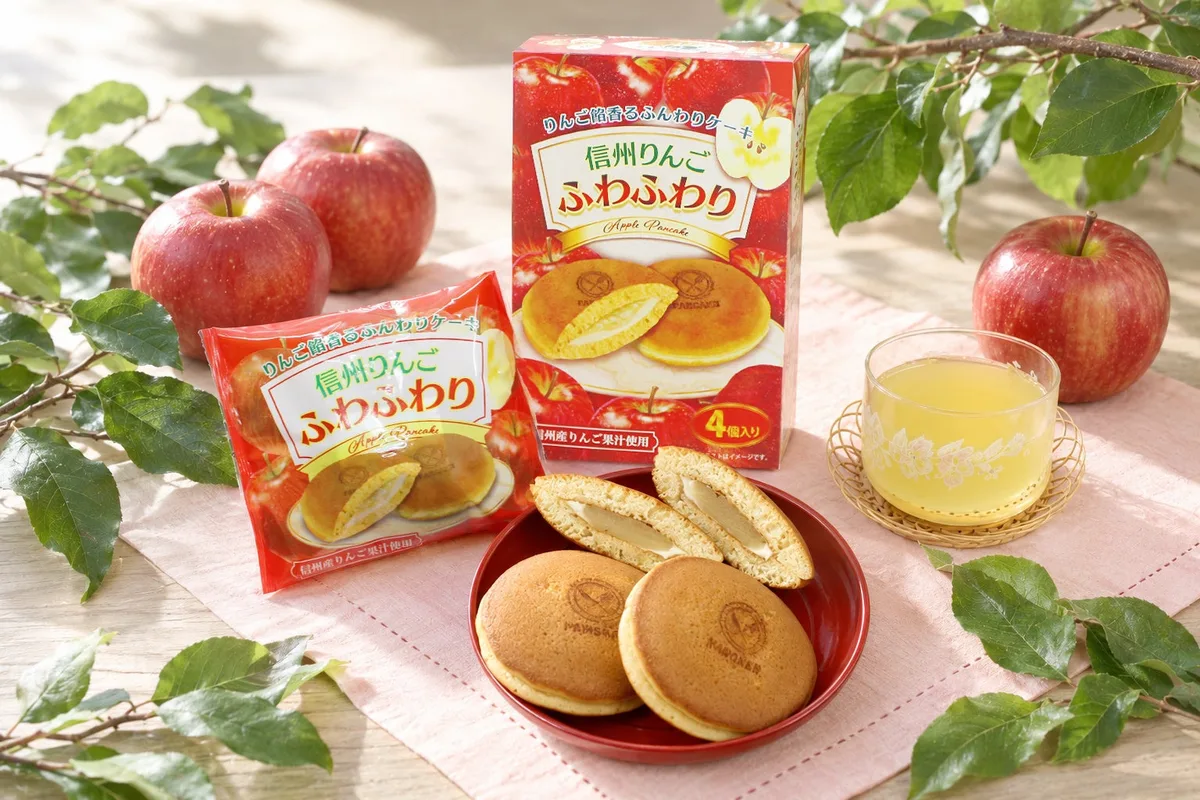赤いパッケージの4個入り箱と1個入り袋、赤い皿に盛られた丸い菓子と切った断面。周りにりんごとりんごの葉、りんごジュースのグラス