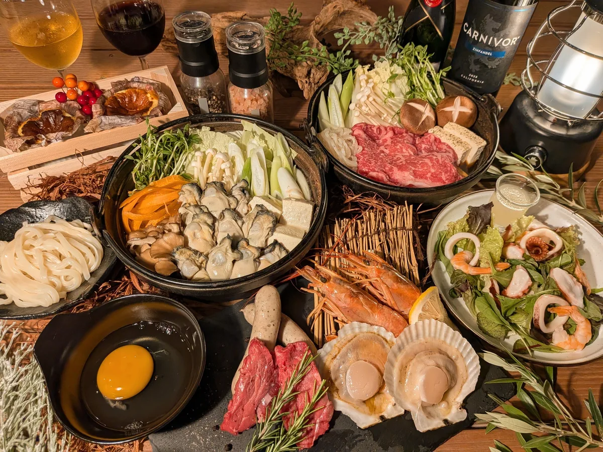 土鍋に並んだ牡蠣と野菜、別の鍋に盛られたすき焼き用の牛肉、海鮮サラダ、うどん、溶き卵、焼き物が木のテーブルに広がる